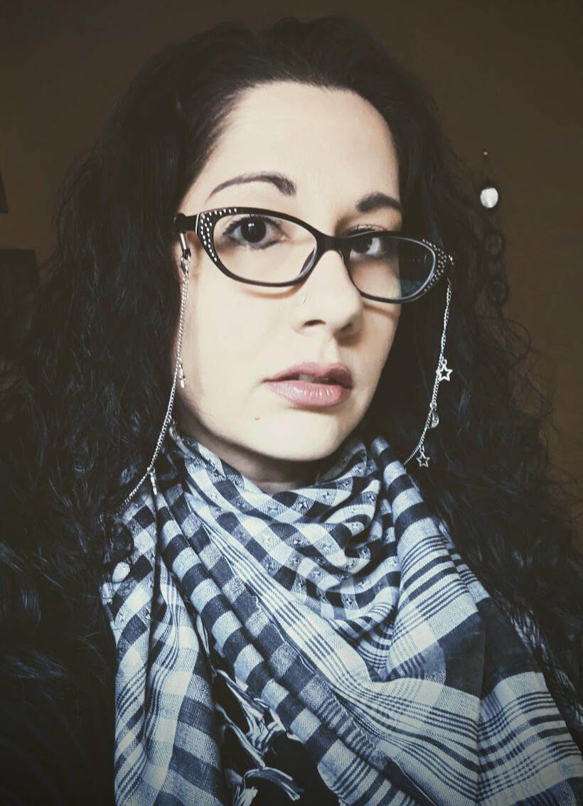 Three-quarter view headshot of Luna, a femme-presenting nonbinary person with long black curly hair and pale skin, wearing a pair of black rhinestone cat's-eye glasses with a silver glasses chain featuring moon and star charms dangling from it. They are al
